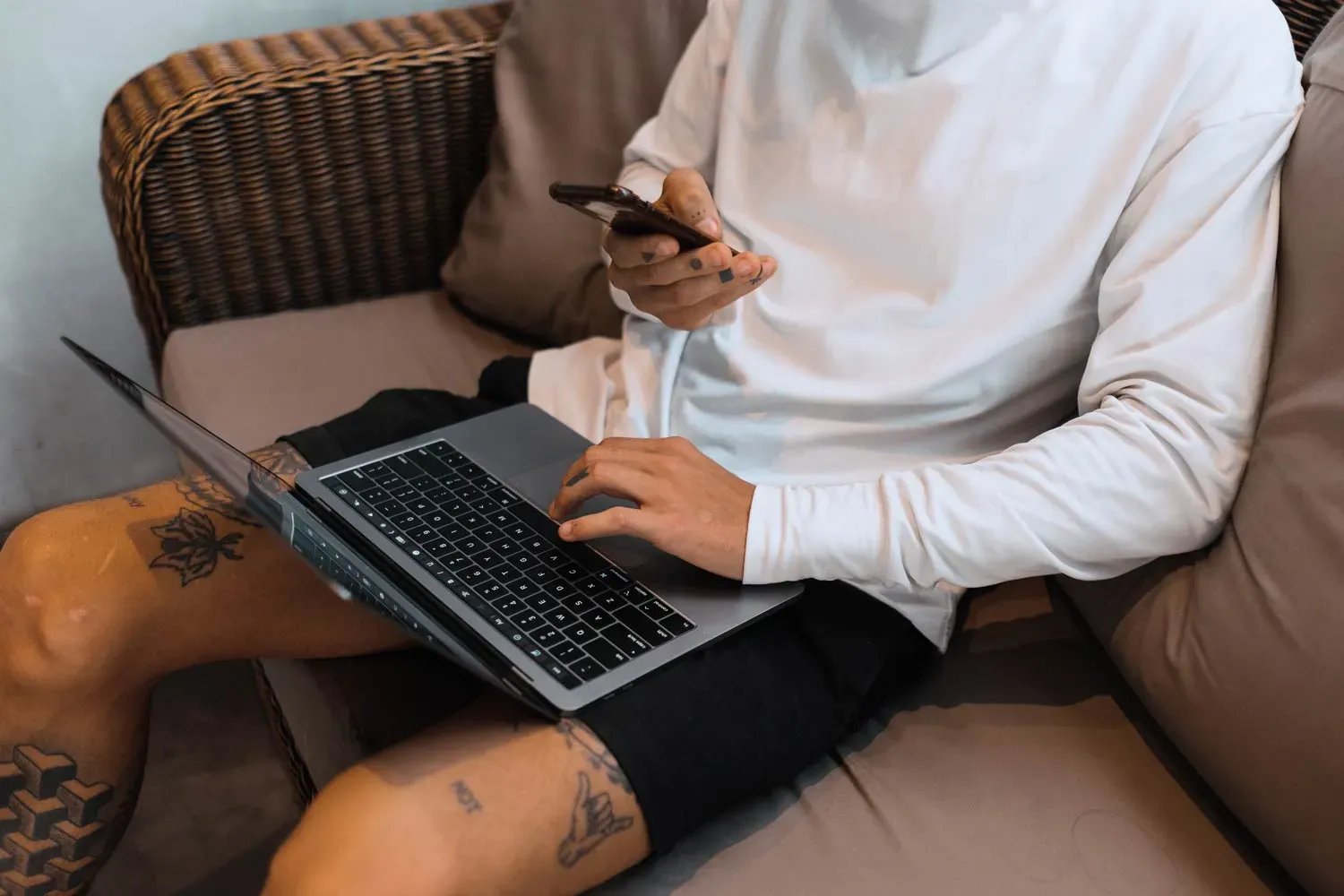 Person sitting on a couch using a laptop and holding a smartphone, wearing a white long-sleeve shirt and black shorts with visible tattoos on legs and hands.