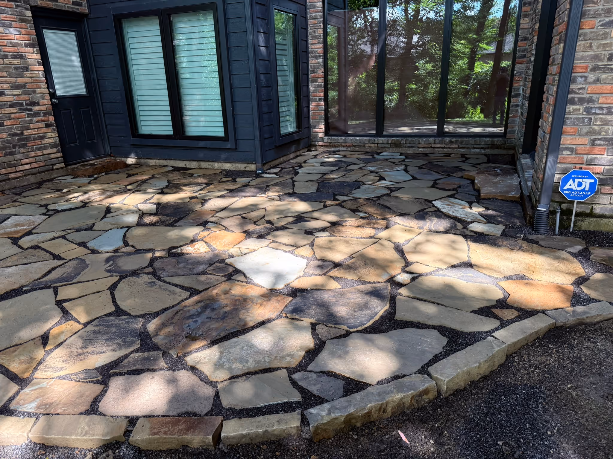 Irregular stone patio with multicolored flagstones in front of a brick and blue-paneled house with a sliding glass door and windows.