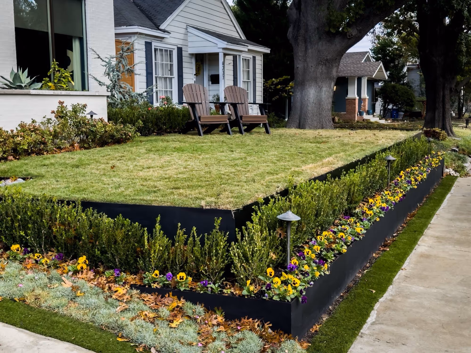 Front yard of a house with raised grass area bordered by black metal edging, flowering plants, shrubs, and two wooden chairs near the entrance.