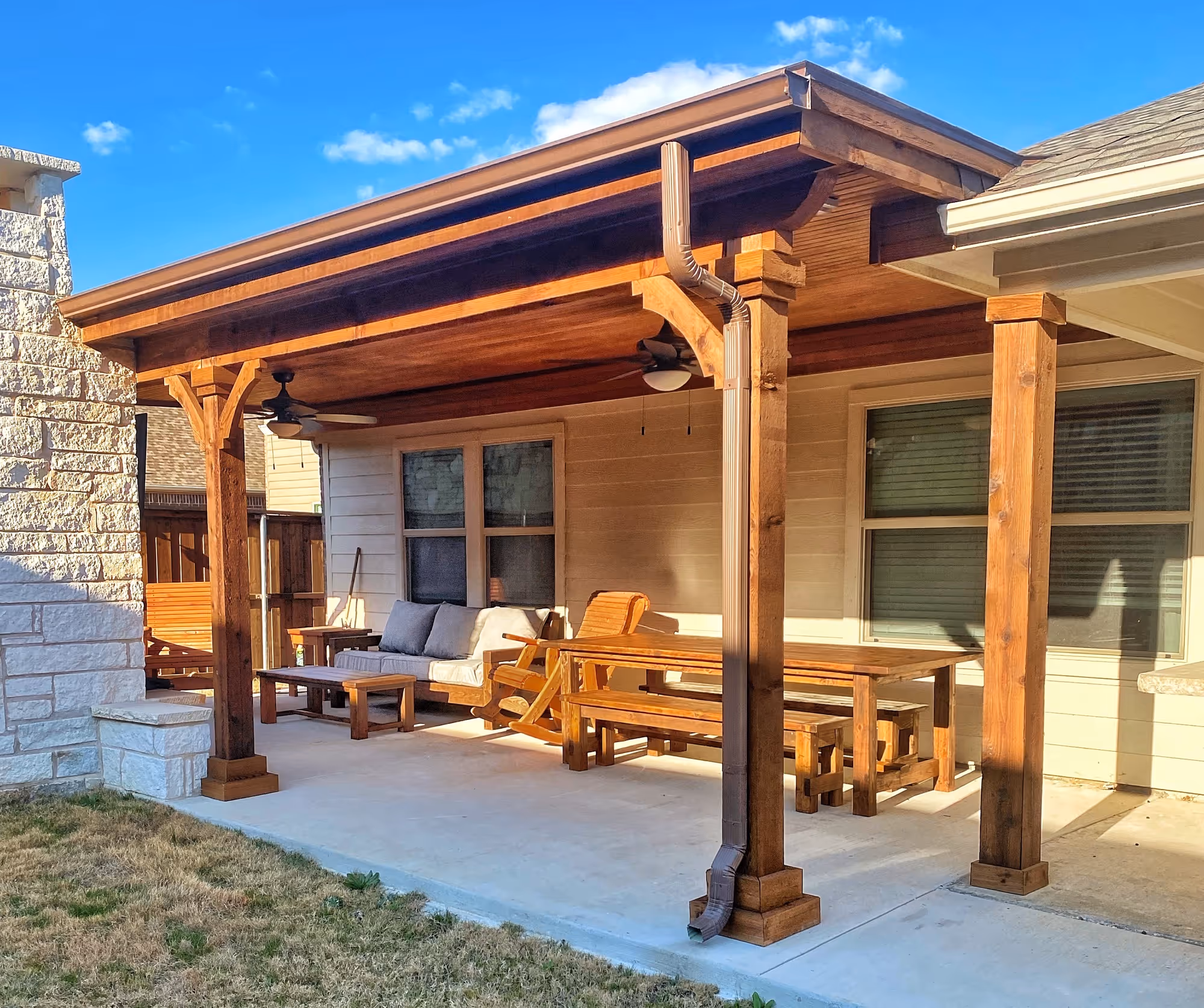 A backyard patio with a wooden pergola overhead, casting shadows on the stone-tiled ground. Benches line the sides of the patio, and a large clay pot sits in the corner. Trees and a fence can be seen in the background.