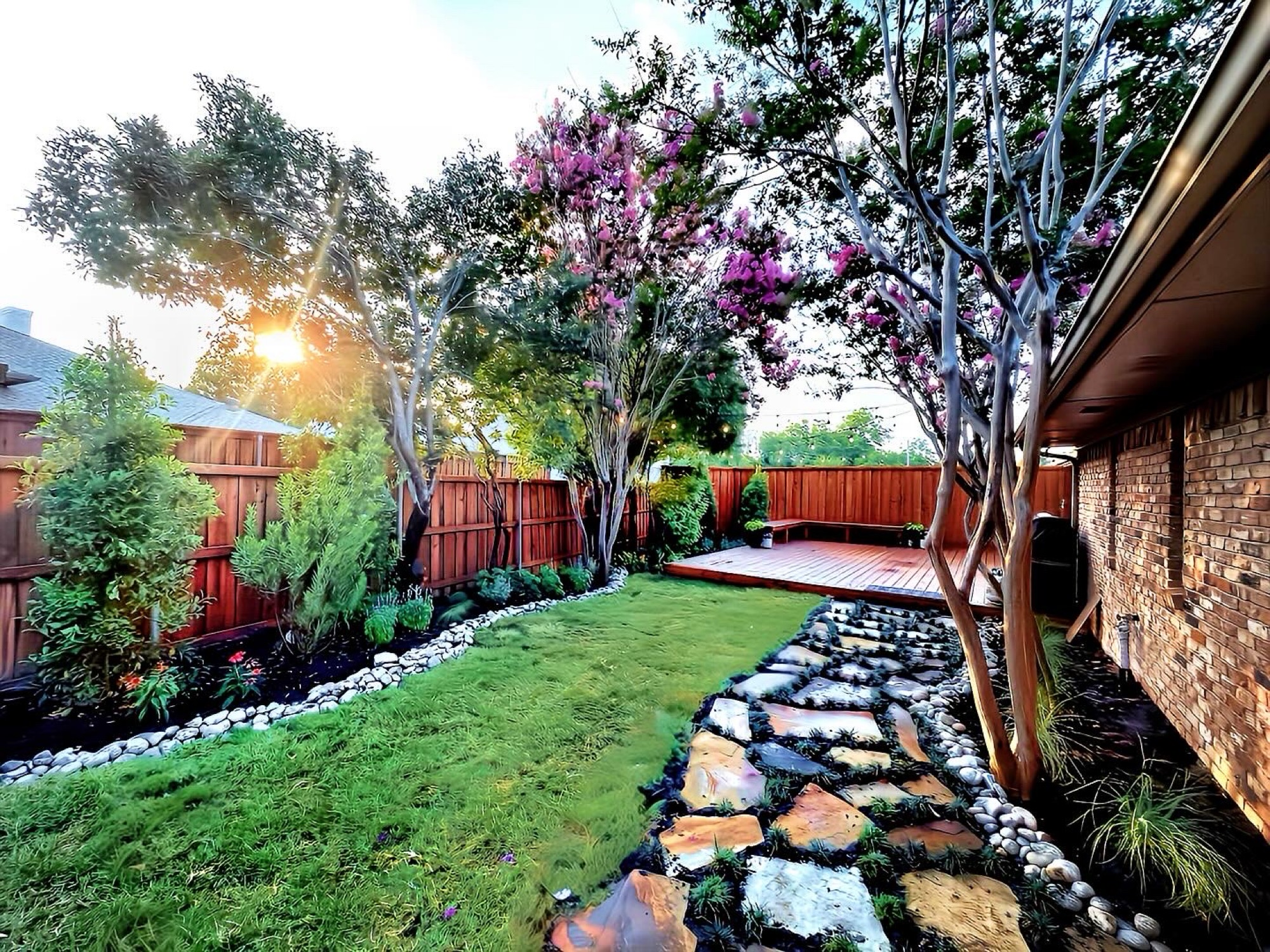 Backyard with green lawn, stone pathway, wooden deck, trees with purple flowers, and a wooden fence at sunset.