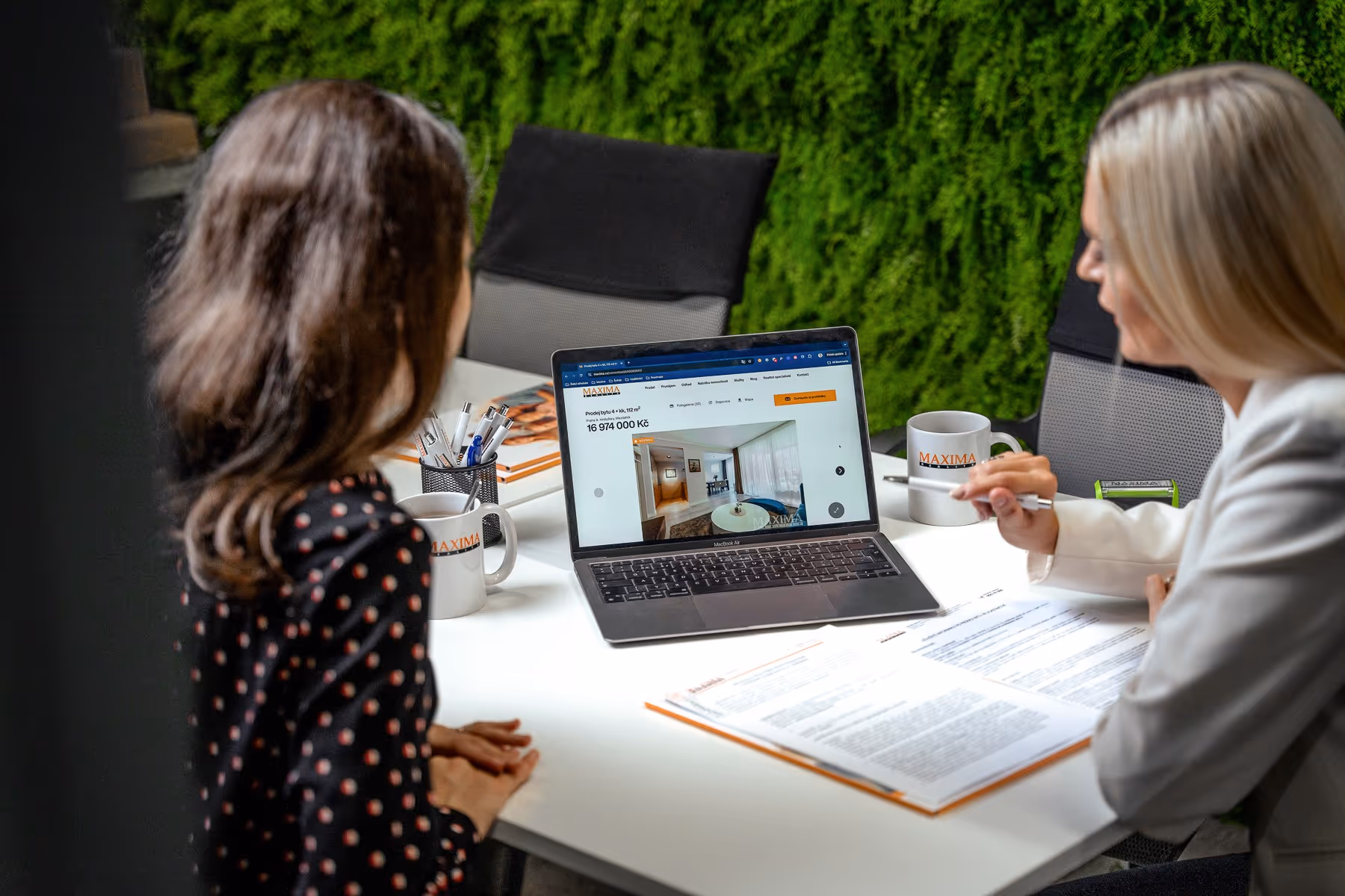 Makléř a zákazník sedí spolu u stolu a počítače. Makléř ukazuje na počítači nemovitost, kterou chce zákazník koupit.