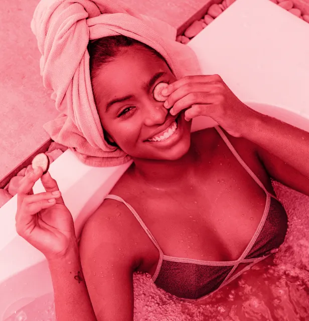 Smiling woman with a towel wrapped around her head, relaxing in a bathtub while holding cucumber slices near her eyes.