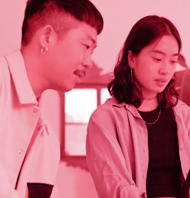 Two young adults looking intently at something, one man with earrings and a woman with shoulder-length hair wearing a necklace.