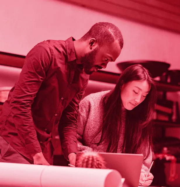 Man and woman collaborating while looking at a laptop, with the woman writing notes.
