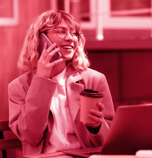 Smiling woman with glasses holding a coffee cup and talking on a smartphone while sitting at a table with a laptop.