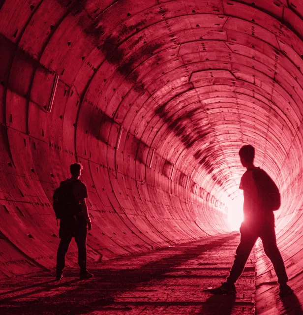 Silhouettes of two people standing inside a curved tunnel illuminated by bright light at the far end.