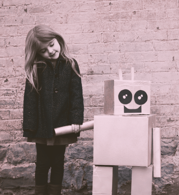 Young girl standing next to a cardboard robot with a smiling face against a brick wall.