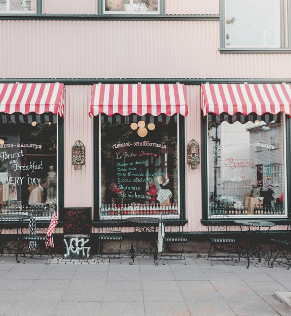 Outdoor seating area with black metal chairs and tables in front of a pink cafe with red-and-white striped awnings and menu items written on the windows.