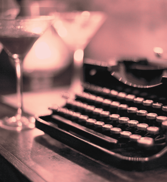 Close-up of a vintage typewriter on a wooden table with two blurred cocktail glasses in the background under soft pink lighting.