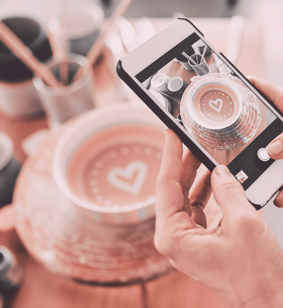 Person holding a smartphone taking a photo of a cup of coffee with a heart design in the foam.