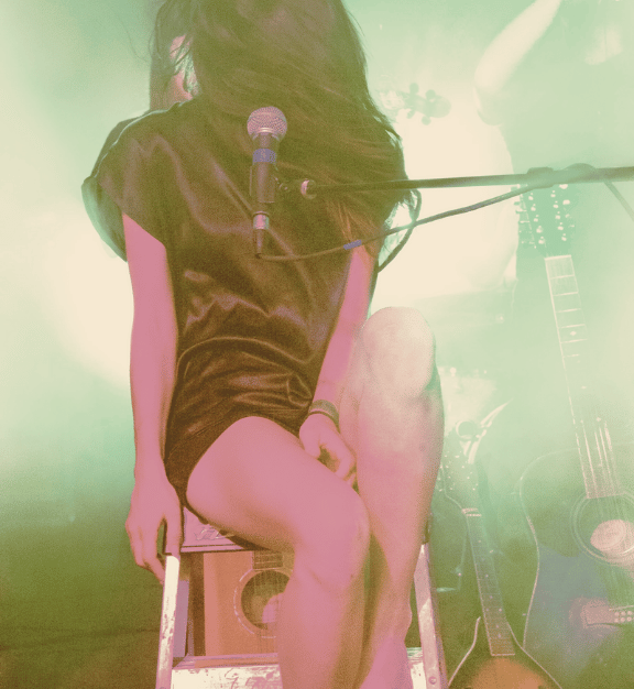 Musician sitting on a stool with a microphone in front and acoustic guitars in the background.