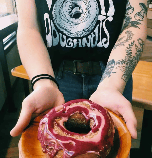 Person with tattooed arms holding a wooden plate with a glazed donut covered in pink and purple icing.