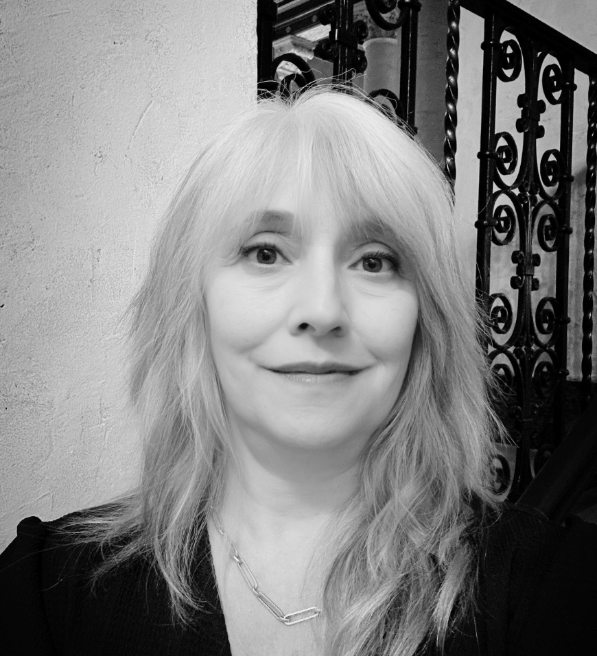 Black and white close-up photo of a woman with light-colored hair smiling slightly, standing in front of a decorative wrought iron railing and a textured wall.