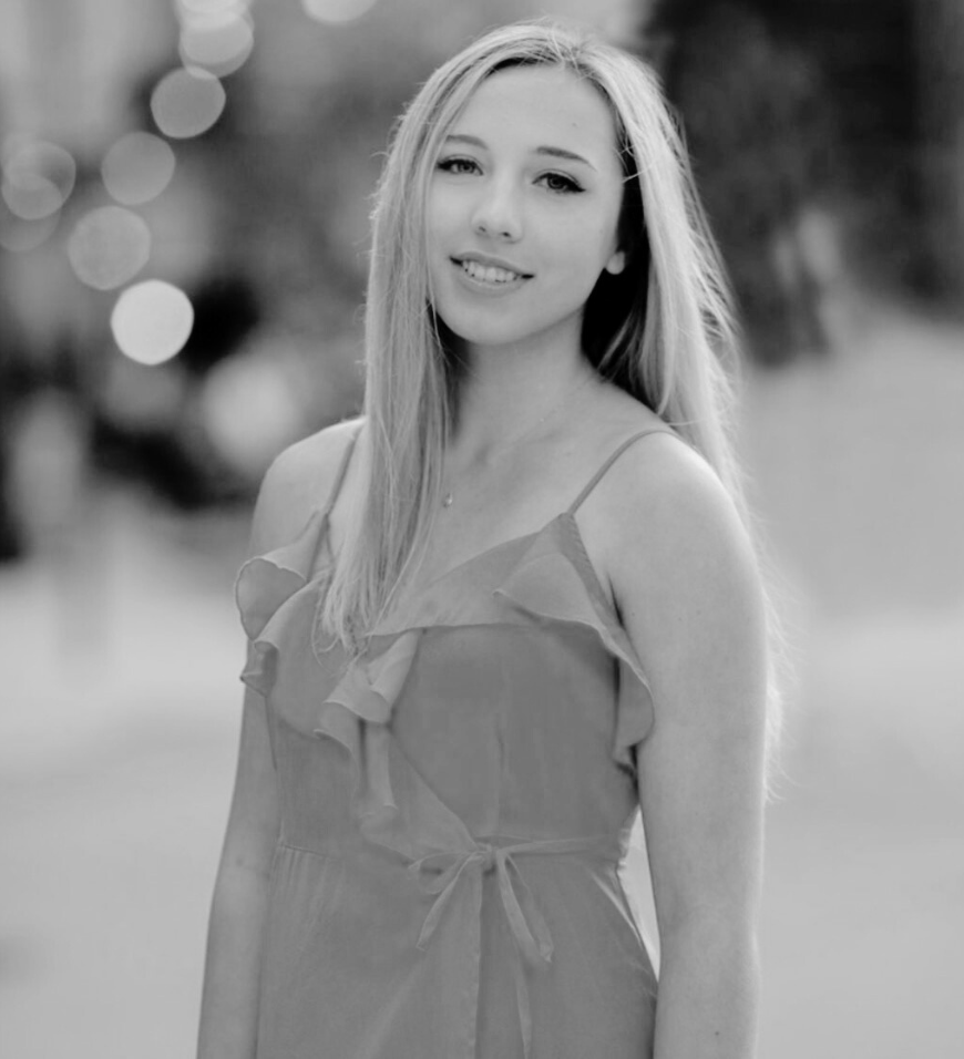 Young woman with long hair smiling, wearing a sleeveless dress with ruffles in an outdoor setting.