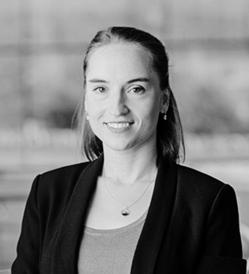 Smiling woman with long hair wearing a dark blazer, a light top, and a pendant necklace.