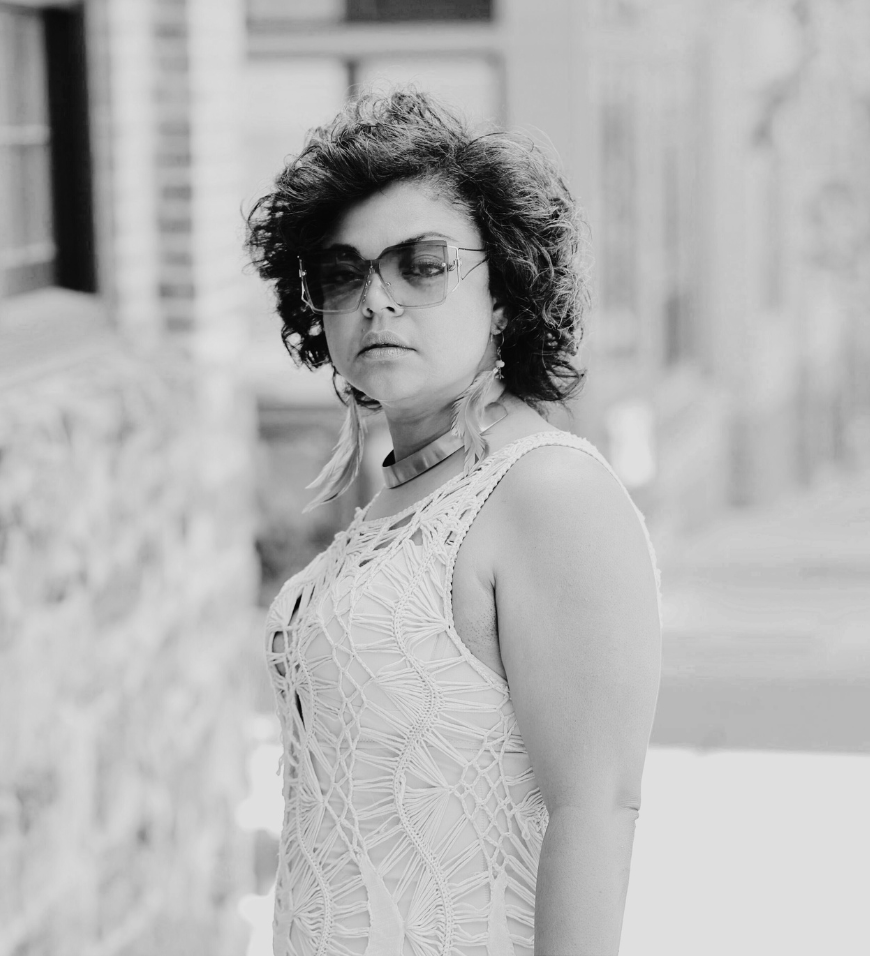 Woman with curly hair wearing large sunglasses, feather earrings, and a sleeveless patterned top.