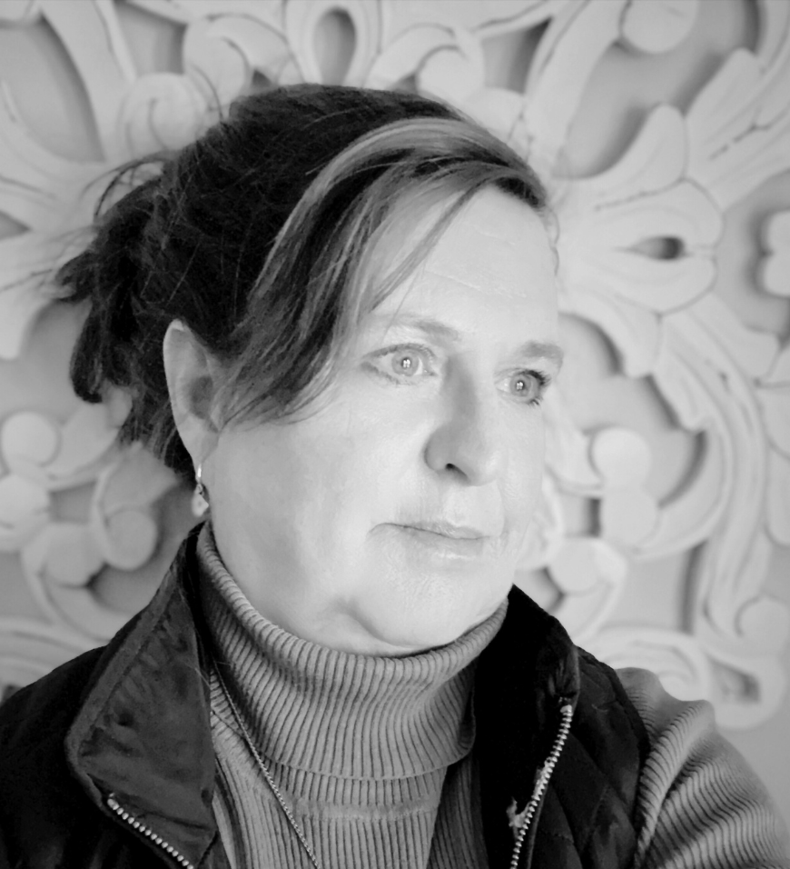 Black and white portrait of a woman with light eyes looking slightly to the right, wearing a turtleneck sweater and a zip-up vest, with her hair tied back and an ornate background.