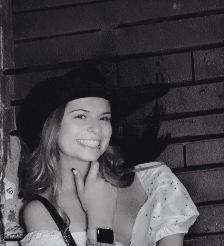 Smiling young woman wearing a wide-brimmed hat and a white off-shoulder blouse, posing with her hand near her chin.