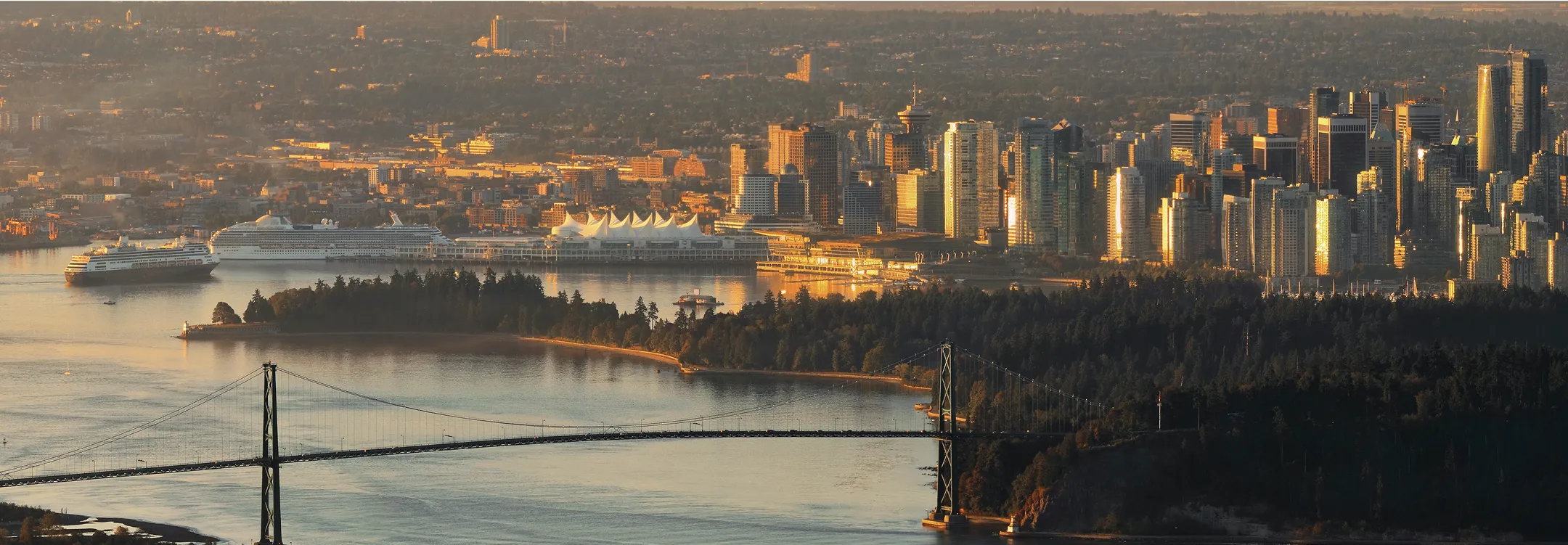 Vancouver's downtown bathed in a warm evening glow, with forested park lands and a historic bridge in the foreground.