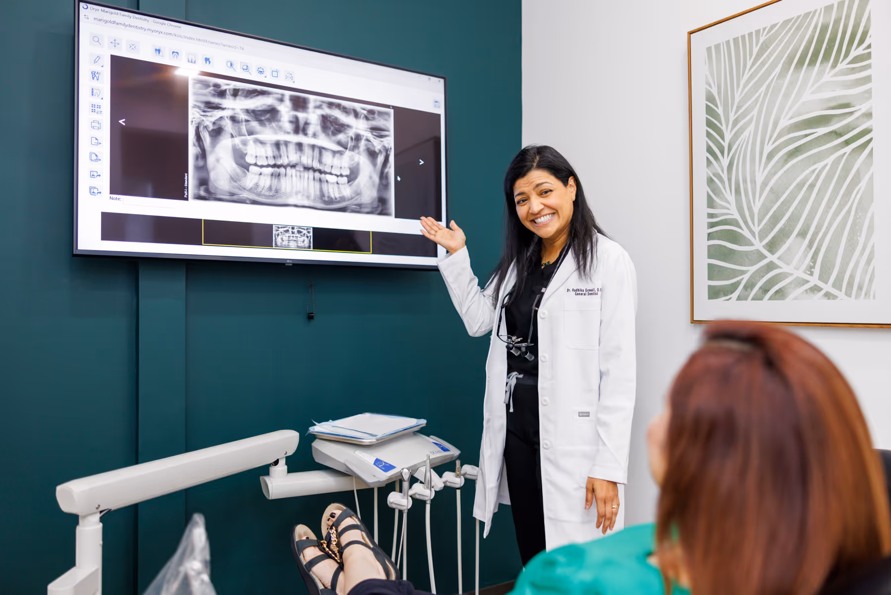 Dentist in a white coat smiling and pointing to a dental X-ray displayed on a screen in a dental office.
