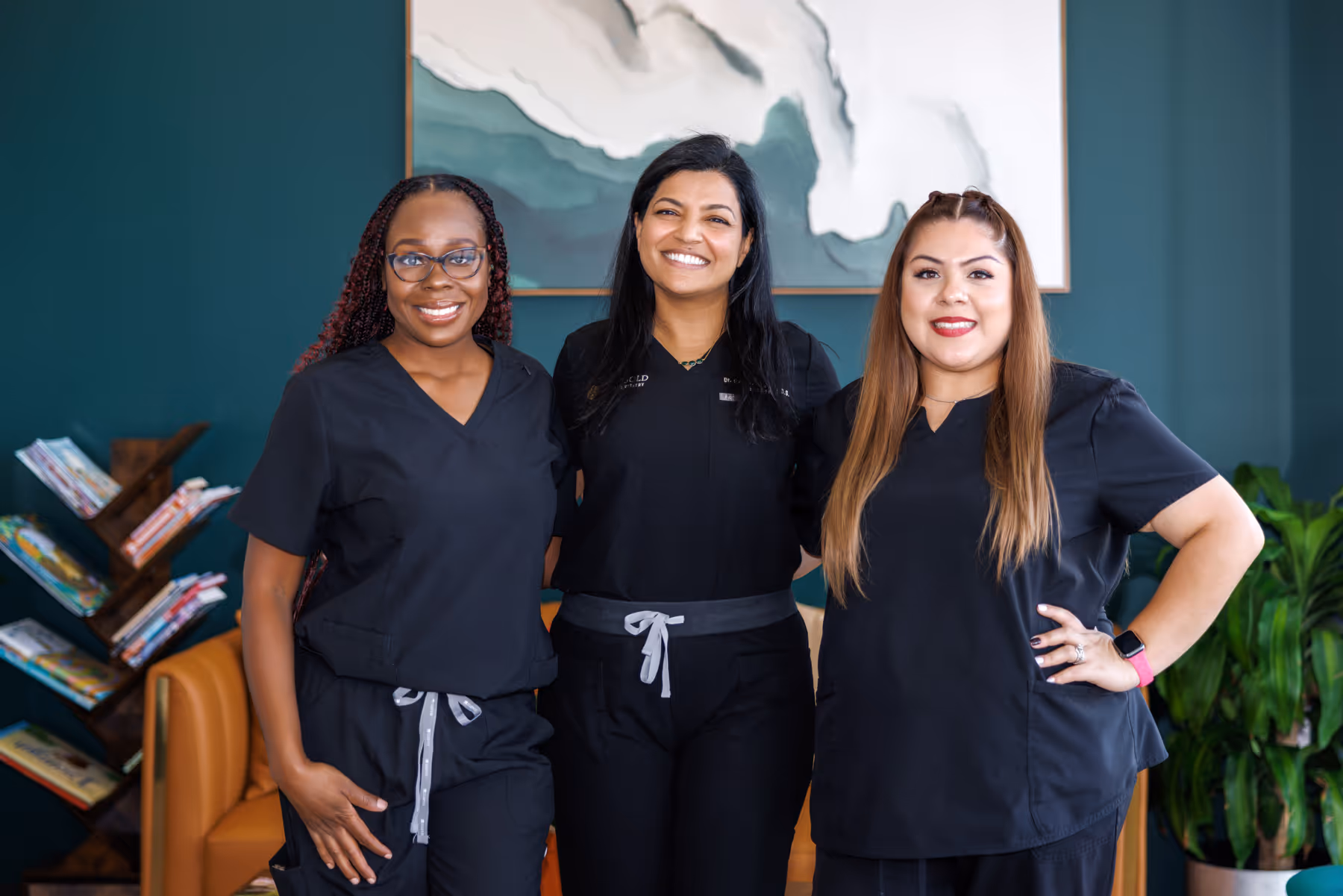 Three smiling women wearing black medical scrubs standing indoors in front of a large abstract painting and a bookcase.