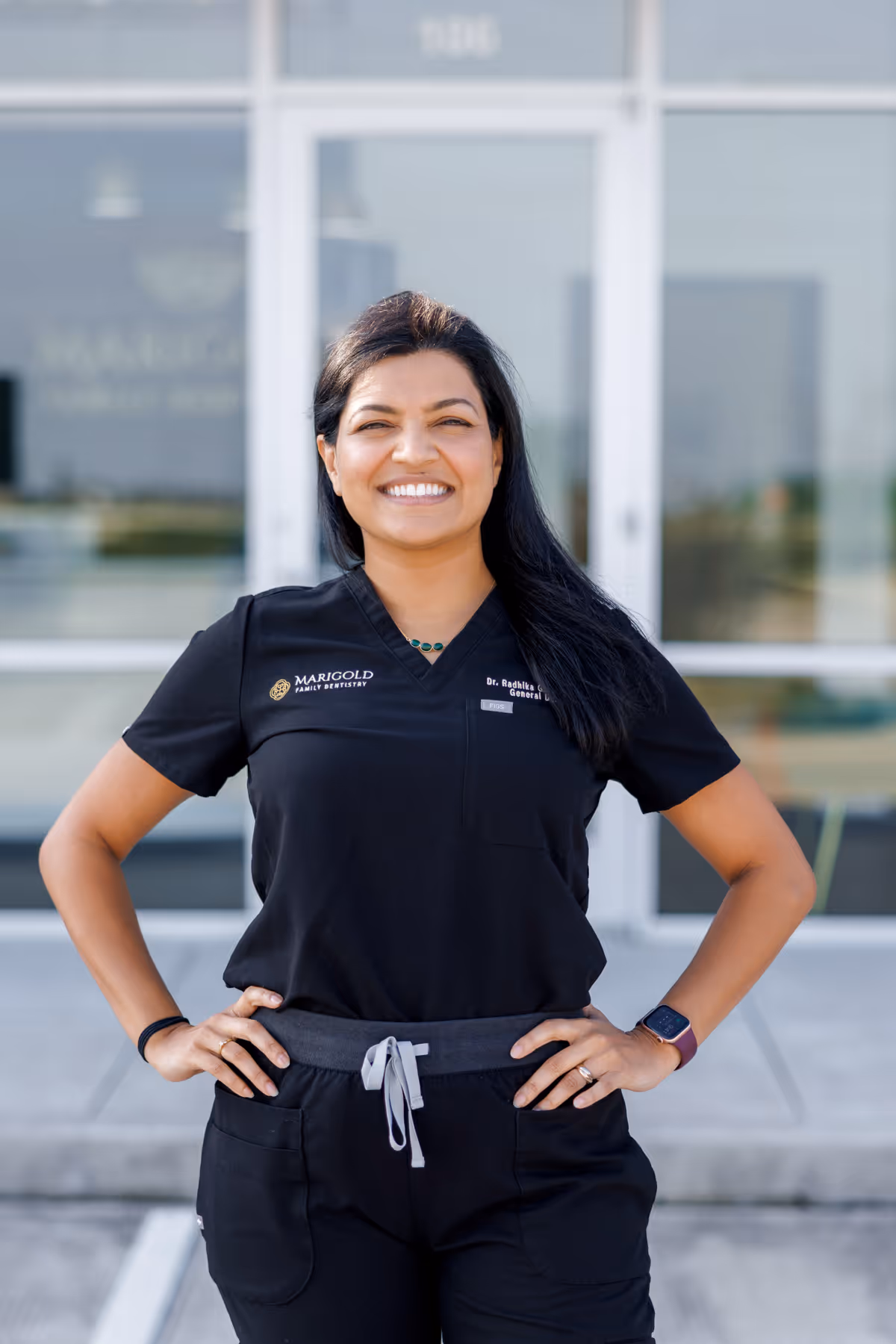 Smiling female dentist in black scrubs standing confidently outside a dental clinic with hands on hips.