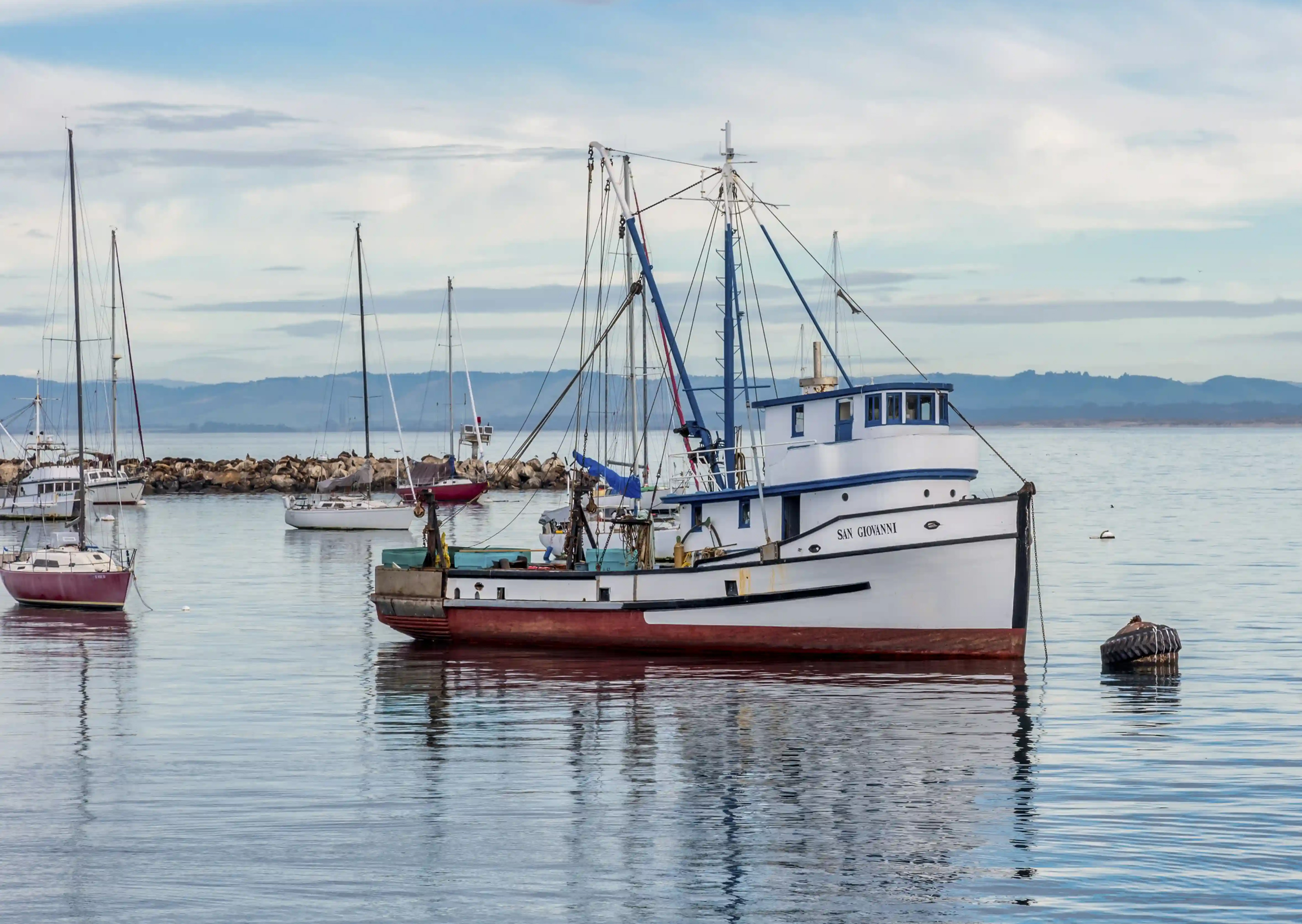 Commercial fishing boat docked in calm harbor waters with masts and shoreline in the background.
