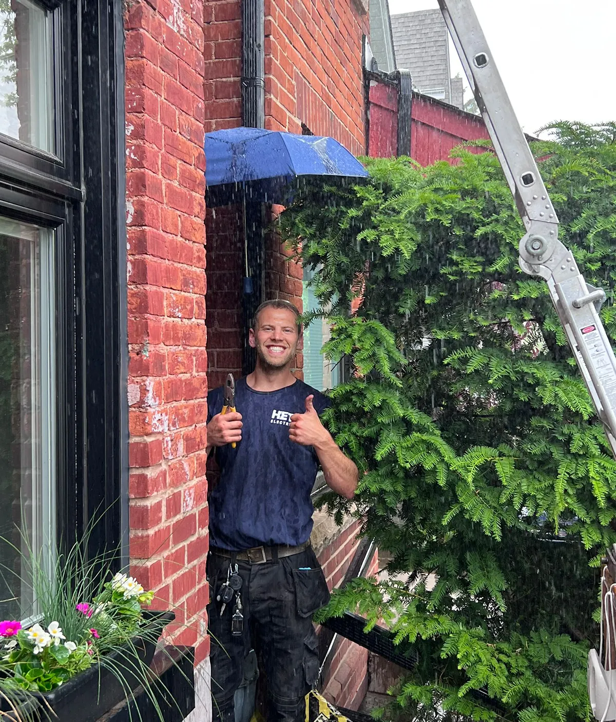 Smiling Hetz Electrical technician giving thumbs up under umbrella in the rain while working outside brick building.