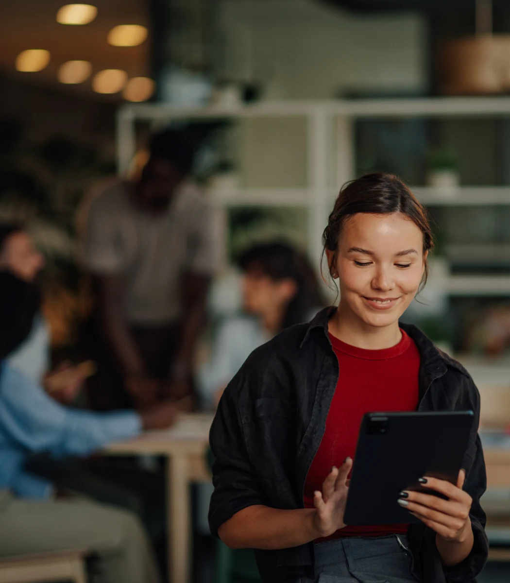Young woman smiling and using a tablet in an office with co-workers in the background.