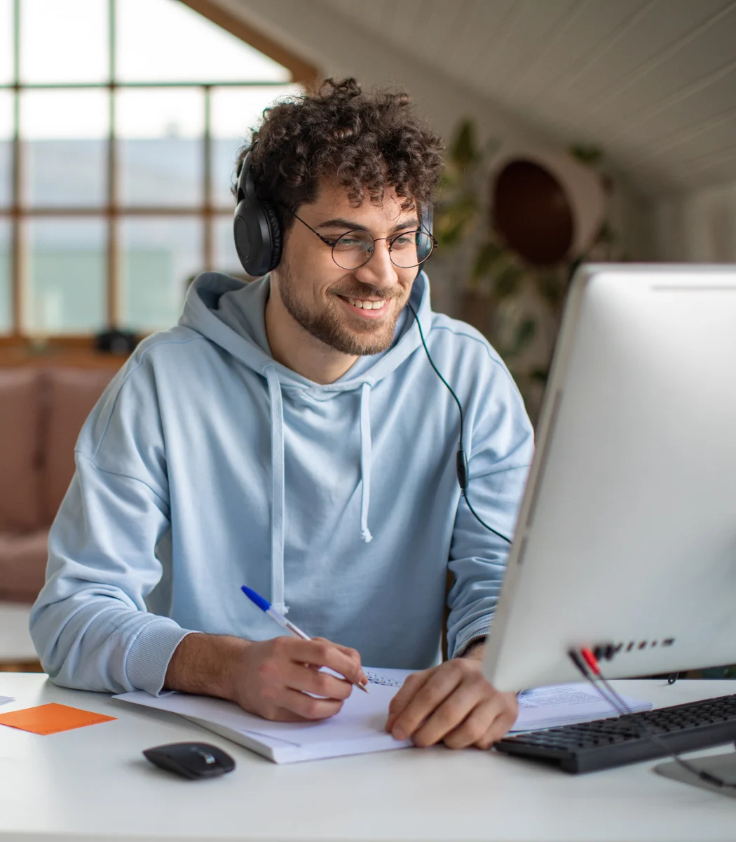 Young man in headphones smiling while writing notes and looking at a desktop computer screen.