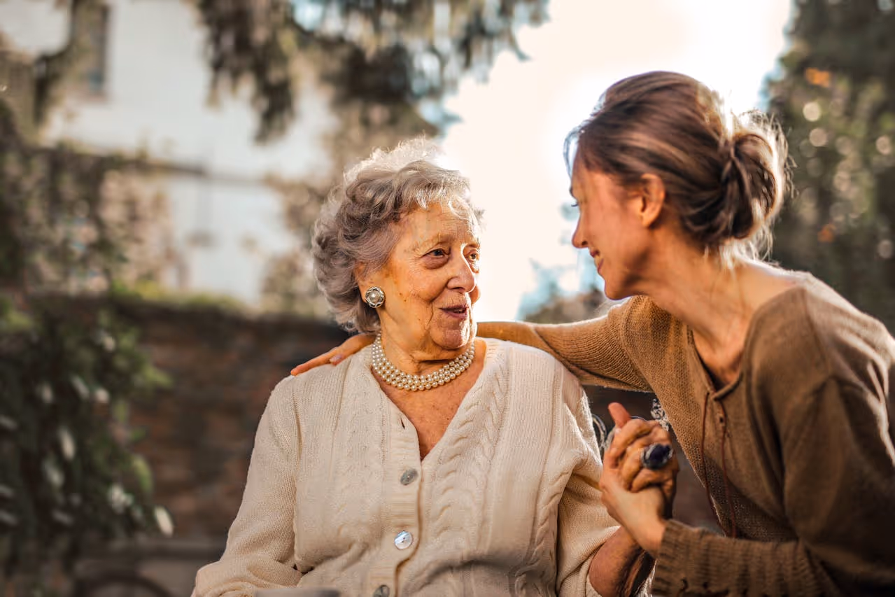 A senior woman smiling warmly while a compassionate caregiver assists her in a comfortable home setting, illustrating the dignity and support of long-term care.