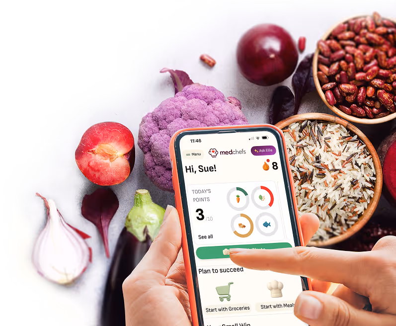 Image of hands holding a phone displaying the MedChefs app homepage. Fruits, vegetables and whole grains in the background