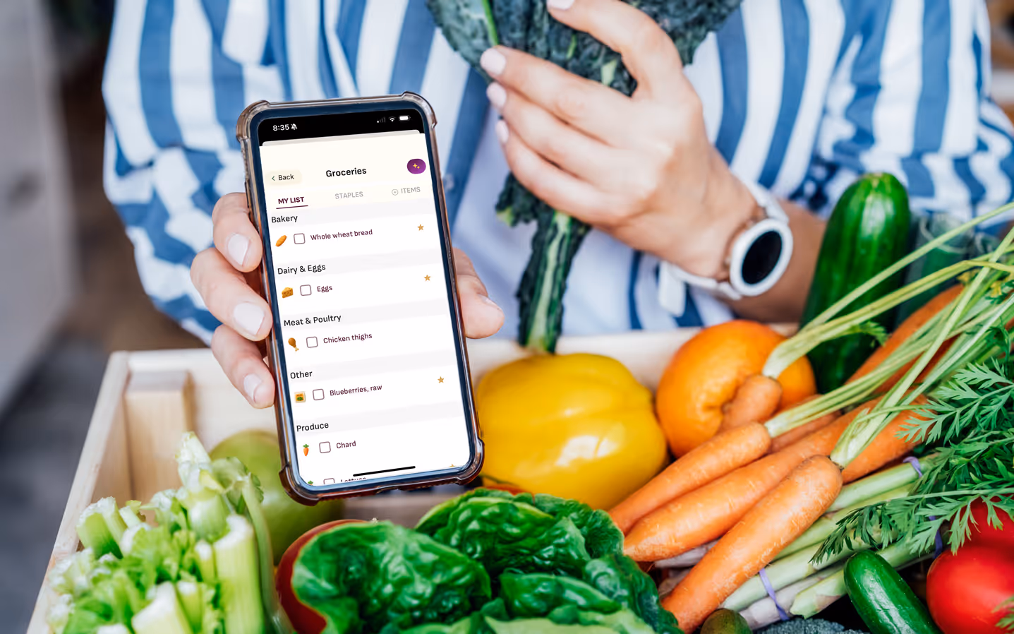 A woman holding a phone showing the Grocery screen on the MedChefs app. A box of fresh vegetables in in the foreground.