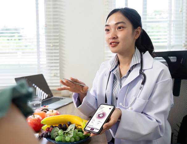 Nutritionist in a white coat showing a healthy eating app on a smartphone to a person across the table with fruits and a laptop in the background.