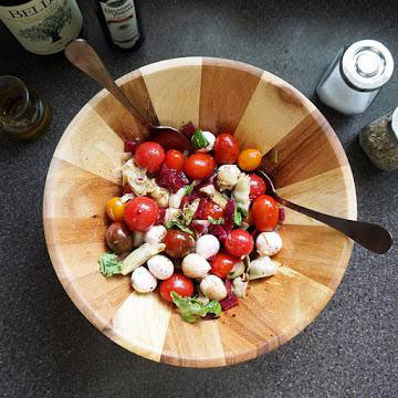 Wooden bowl with beet and artichoke salad including cherry tomatoes, mushrooms, and leafy greens with two spoons.