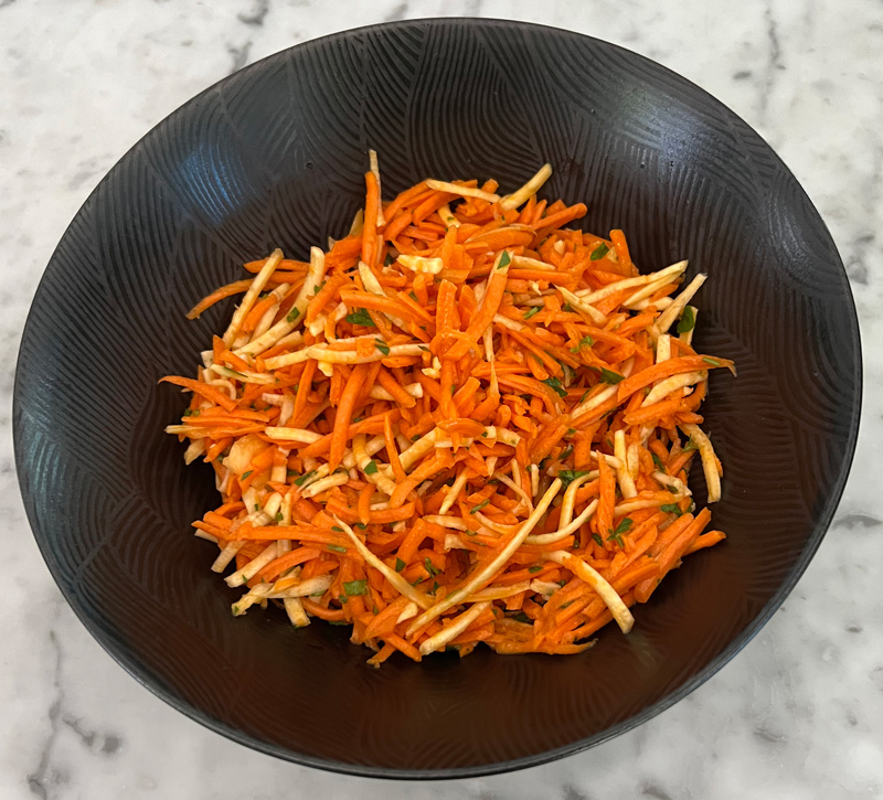 Black bowl filled with shredded carrot and celery root salad with herbs on a marble surface.