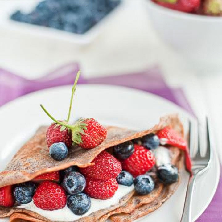 Buckwheat crepe filled with ricotta cheese and topped with fresh strawberries and blueberries on a white plate with a fork.
