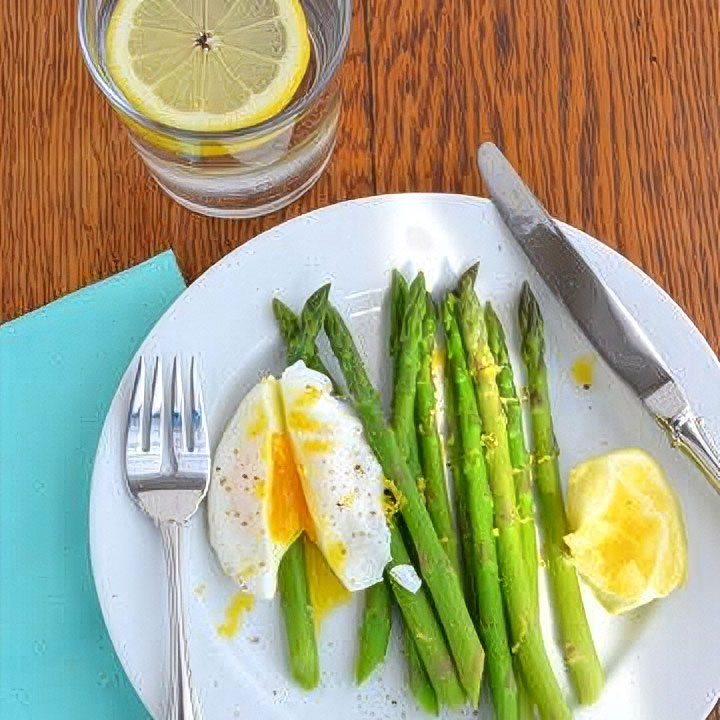 Plate with poached eggs on asparagus, lemon wedge, fork, and knife, next to a glass of water with lemon slice on a wooden table.