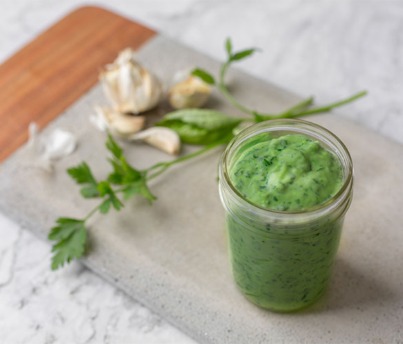 Clear jar filled with green herb sauce placed on a cutting board with garlic and parsley in the background.