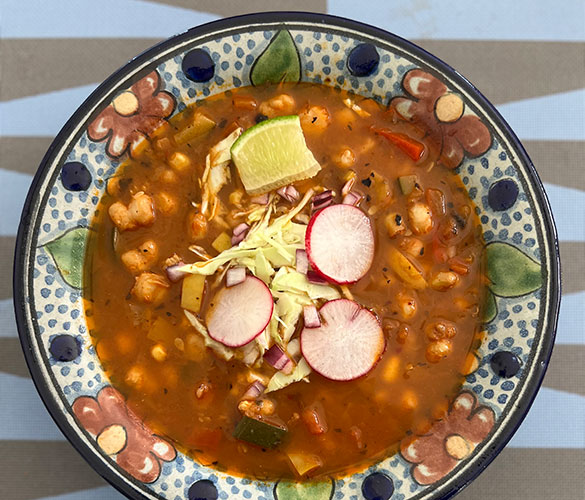 Bowl of vegetarian pozole soup garnished with lime wedge, sliced radishes, shredded cabbage, and diced onions.