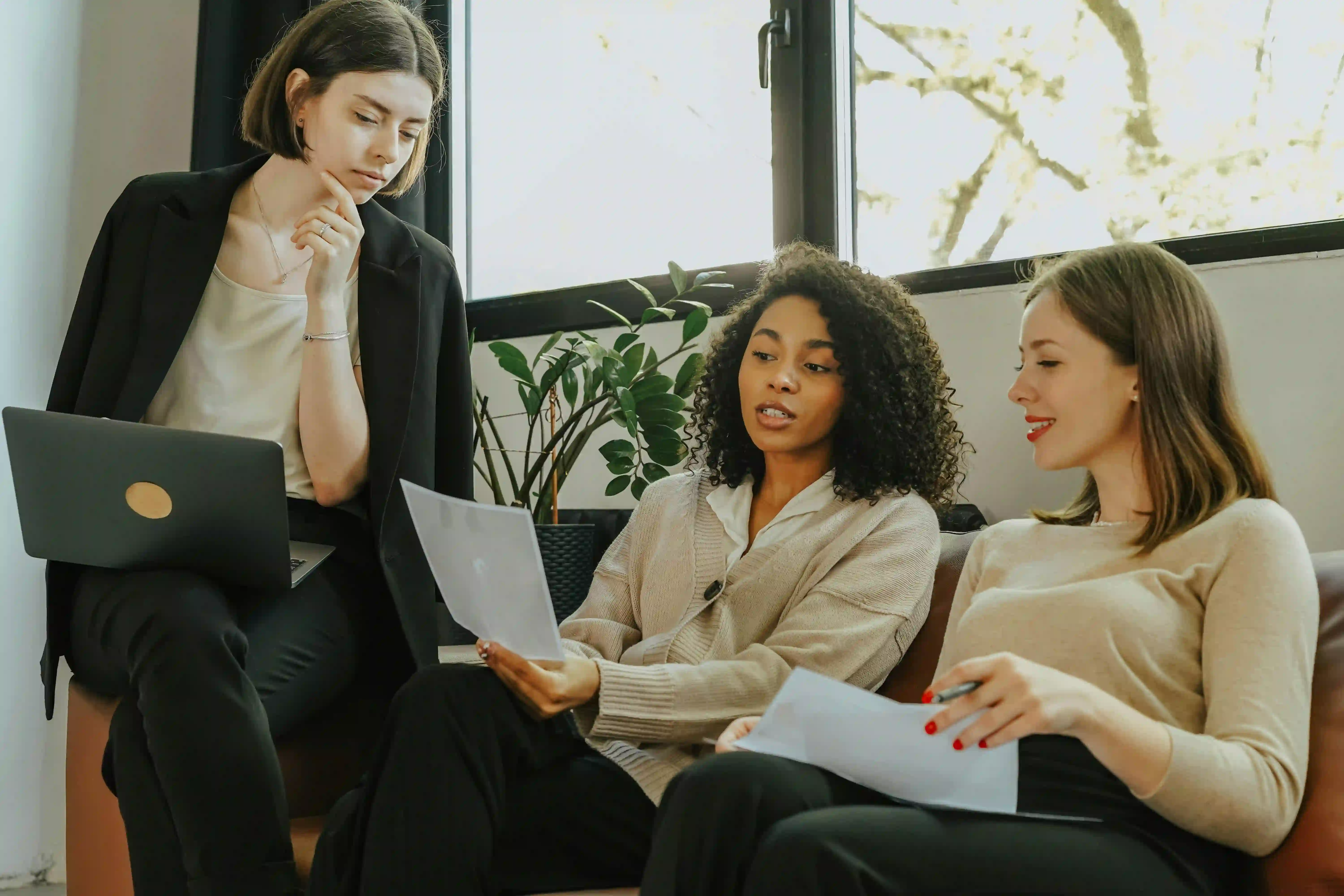 Three female business professionals working together informally at a couch in an office, collectively reviewing a printed document.