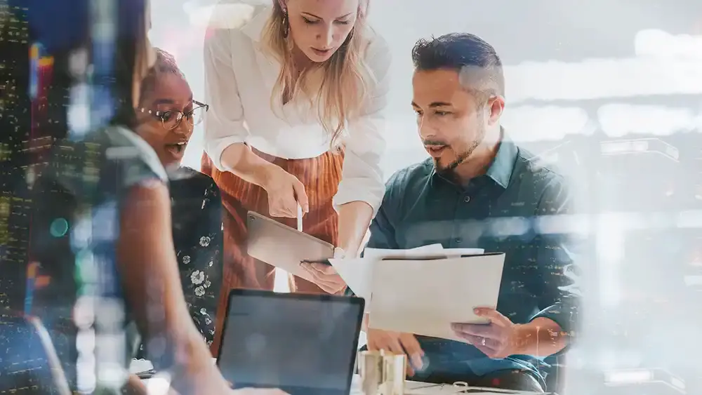 Four young business professionals in a modern-looking office with three of them glancing at a tablet.
