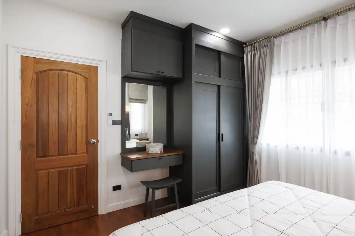 Modern bedroom with a floor-to-ceiling black wardrobe, integrated vanity, and a teak door.