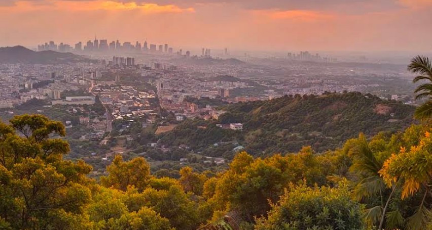 Panoramic view of Chiang Mai city at sunset from Doi Suthep mountain, with warm amber and orange sky, lush tropical trees in the foreground and the city skyline stretching across the valley below.