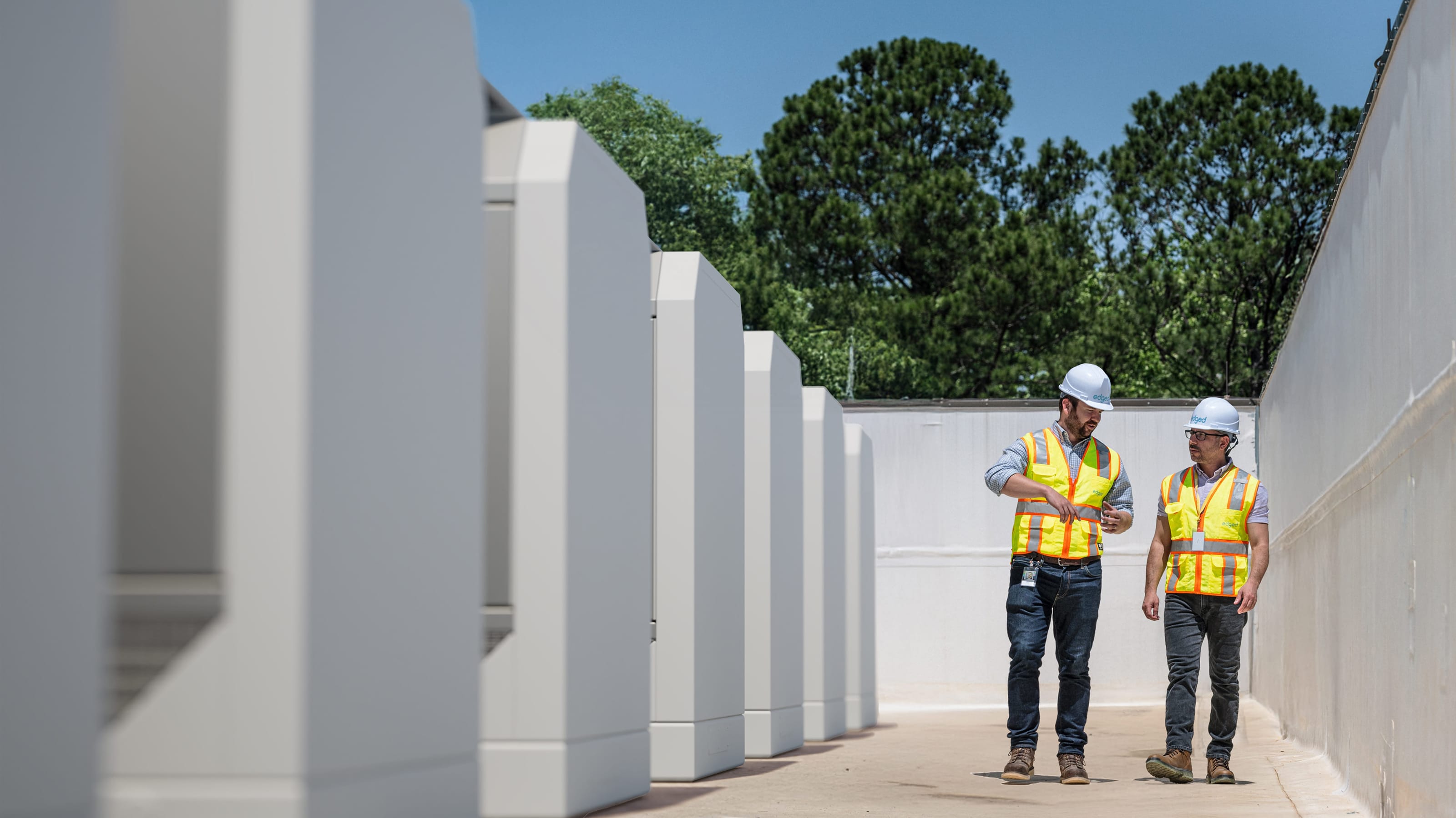 Two construction workers wearing hard hats and orange safety vests walk and talk beside a row of large white industrial equipment outdoors.
