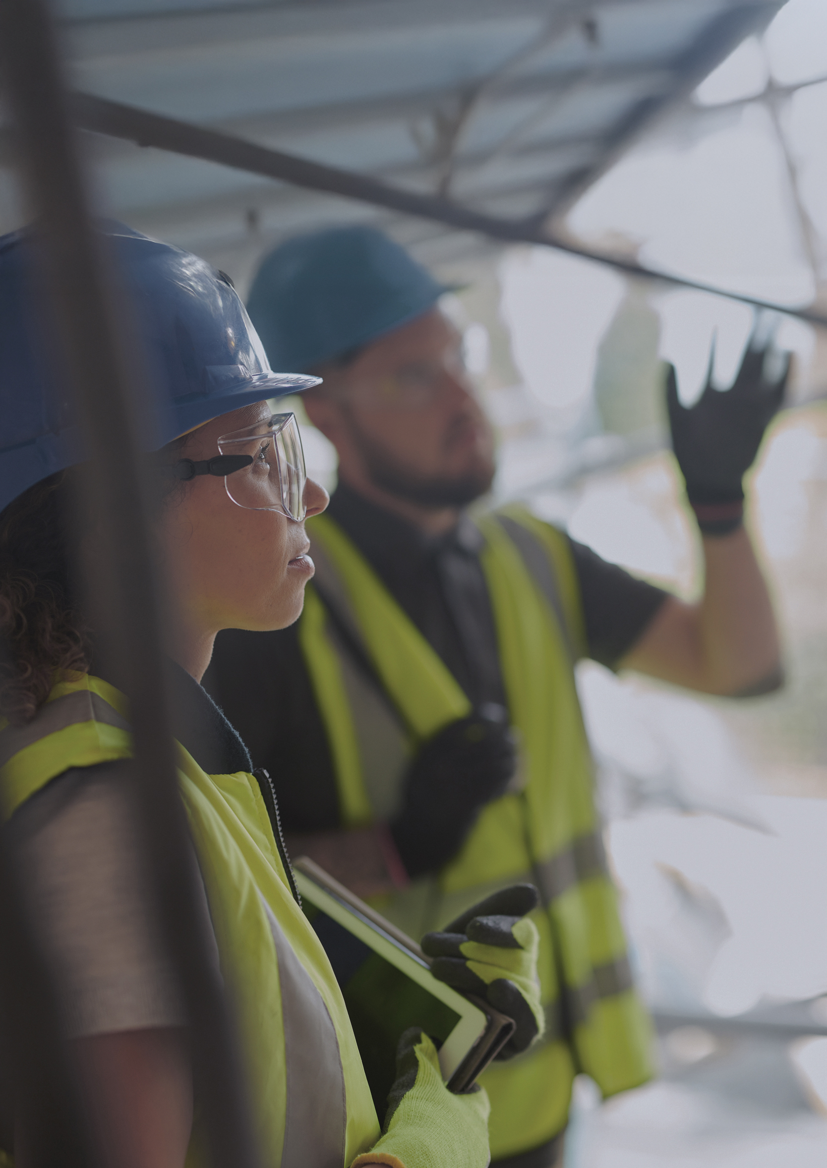 Two construction workers wearing blue helmets and yellow safety vests, one holding a tablet and the other gesturing with a gloved hand.
