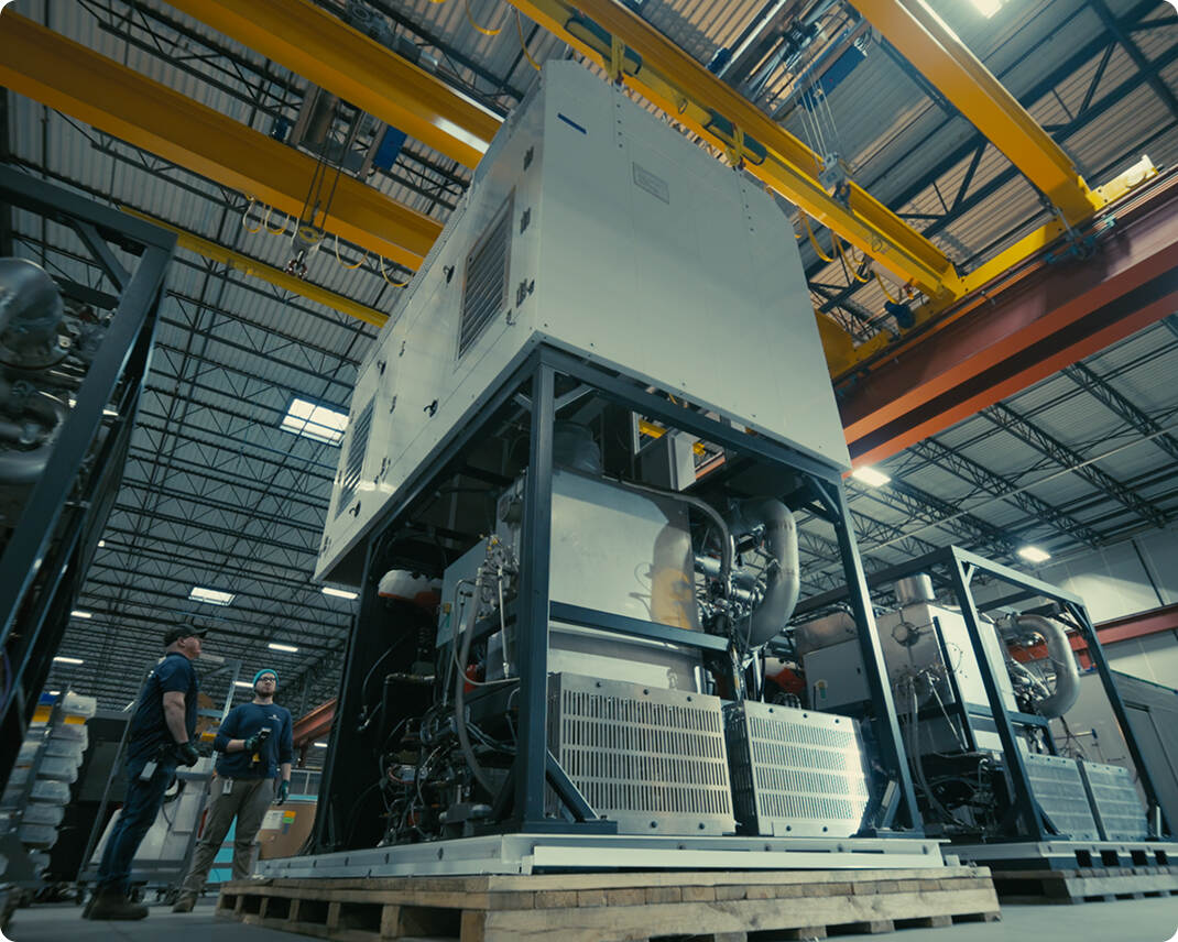 Two workers in a factory inspecting large industrial machinery units mounted on wooden pallets under yellow overhead cranes.