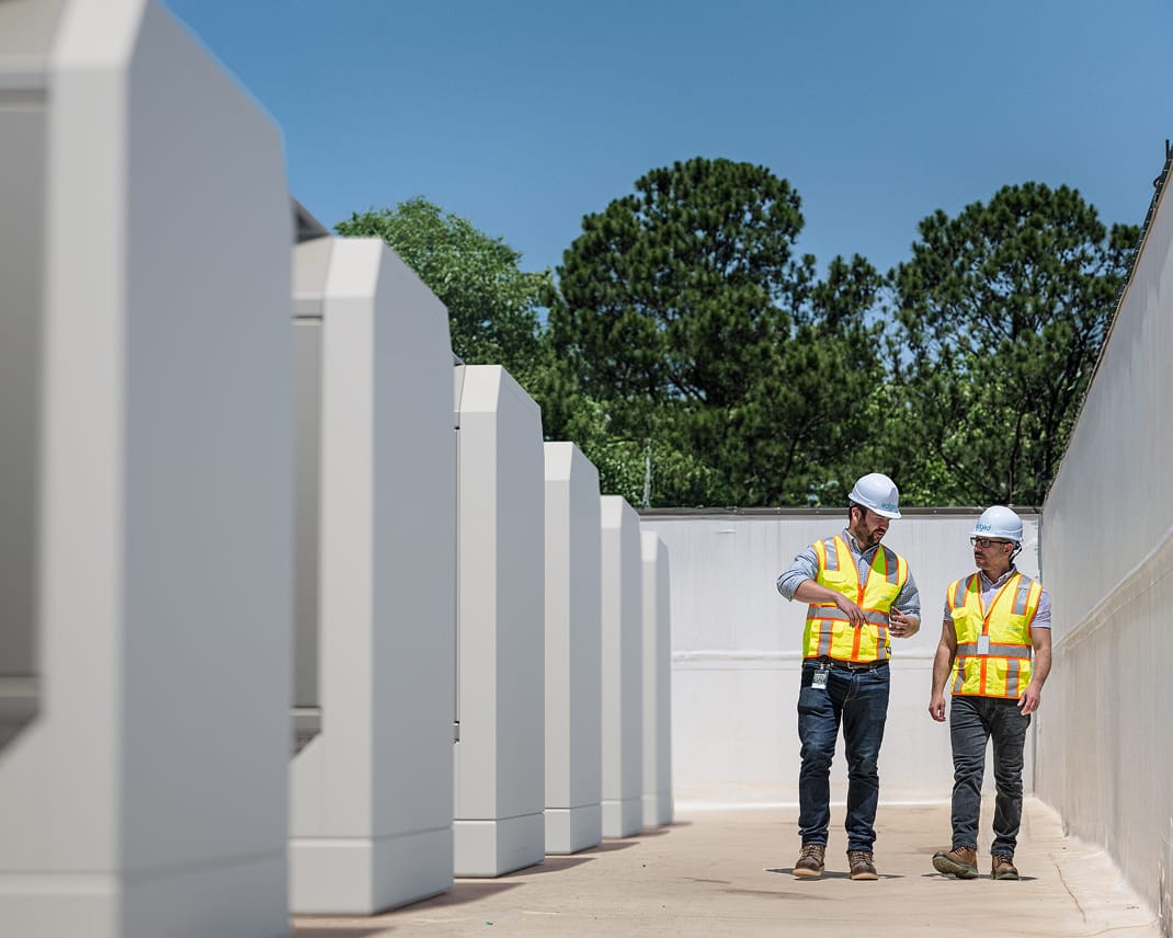 Two construction workers wearing helmets and reflective vests walking between large industrial equipment units outdoors under clear blue sky.