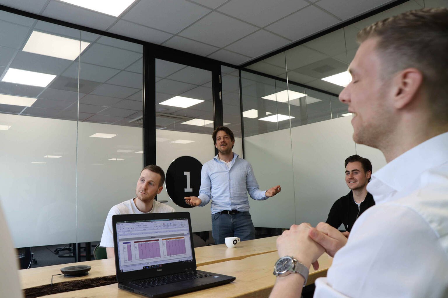 Vier groeileaders in een vergaderruimte; één staat en spreekt terwijl de anderen aan tafel zitten en luisteren, met een opengeklapte laptop op tafel.